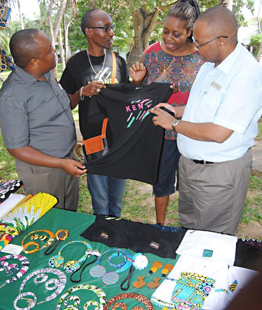 An artist from Rwanda,Ruyange Jose [2nd left] shows a T- Shirt to General Manager of' Serena Beach Resort and Spa in Mombasa, Herman Mwasaghua [right] during Mombasa Farmers and Artisans Market held at the Hotel. 