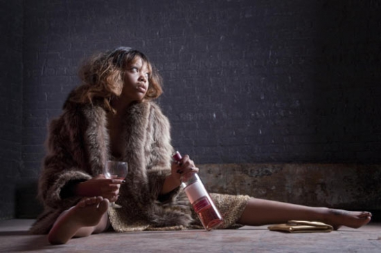 Woman in evening wear and fur coat sits sprawled on the floor with a bottle of wine